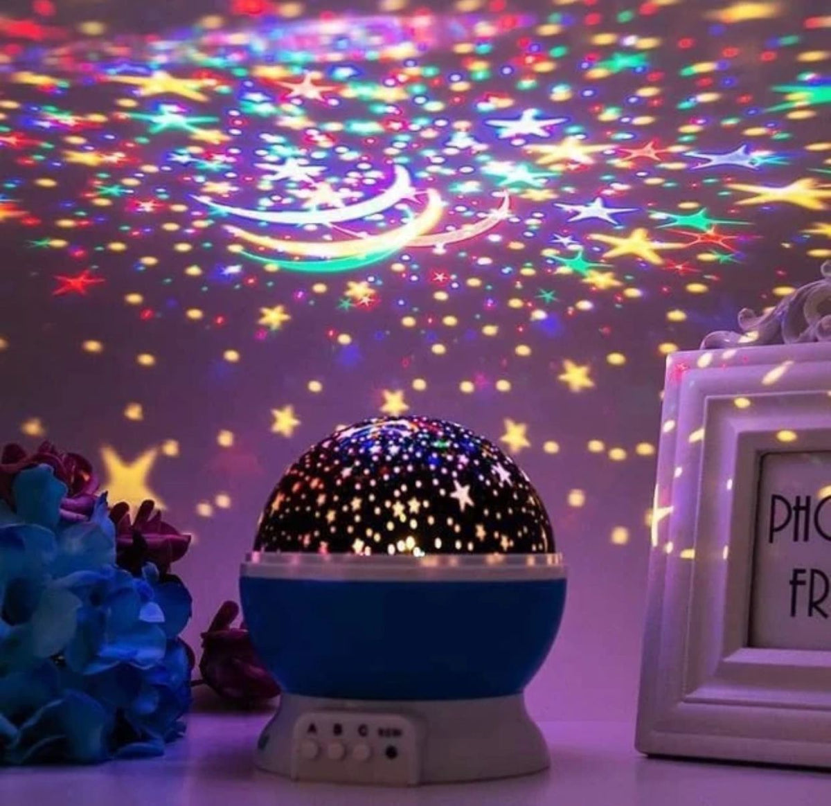 An egg-shaped rose gold constellation projector lamp resting on a blue surface, casting vibrant star patterns and constellations onto a deep blue wall, flanked by a gold armillary sphere model, a stack of vintage books with a small unicorn figurine, and a dreamcatcher.