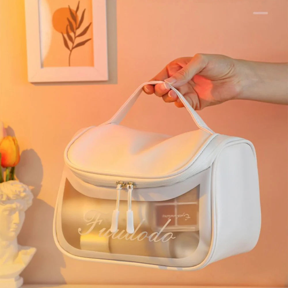 White travel makeup bag featuring a clear window panel and top handle being held against a warm background.